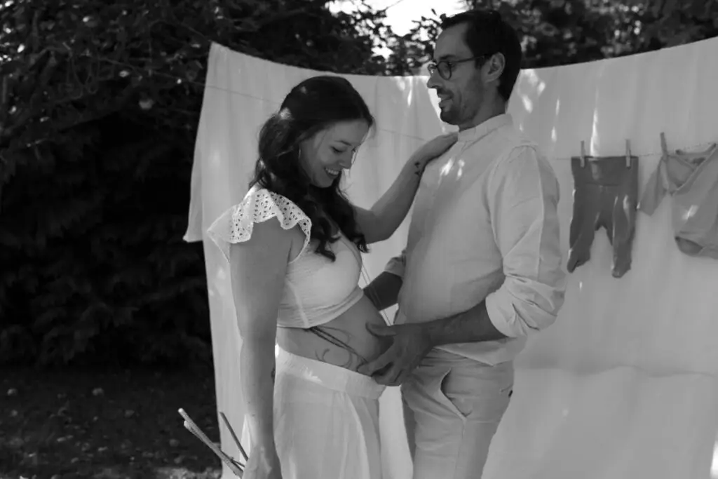 Pregnant woman and partner outdoors, smiling beside a white sheet hanging on a clothesline.